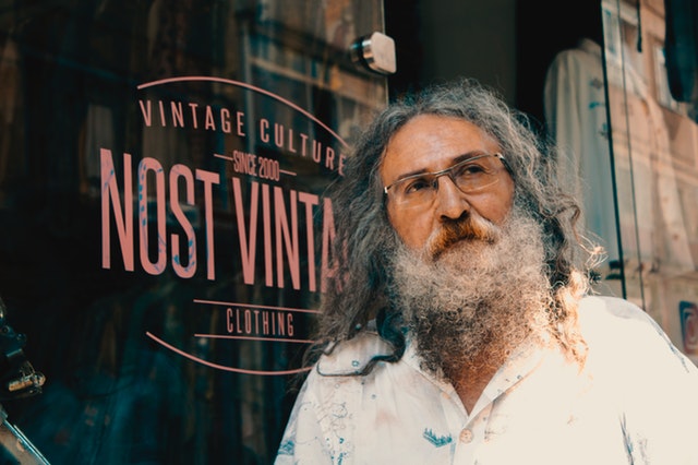 Starší muž s dlhou bradou a vlasmi stojí pred skleneným výkladom.jpg
